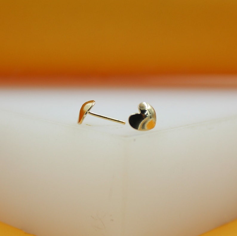 A pair of glossy gold heart-shaped stud earrings rests on a white surface against an orange backdrop.