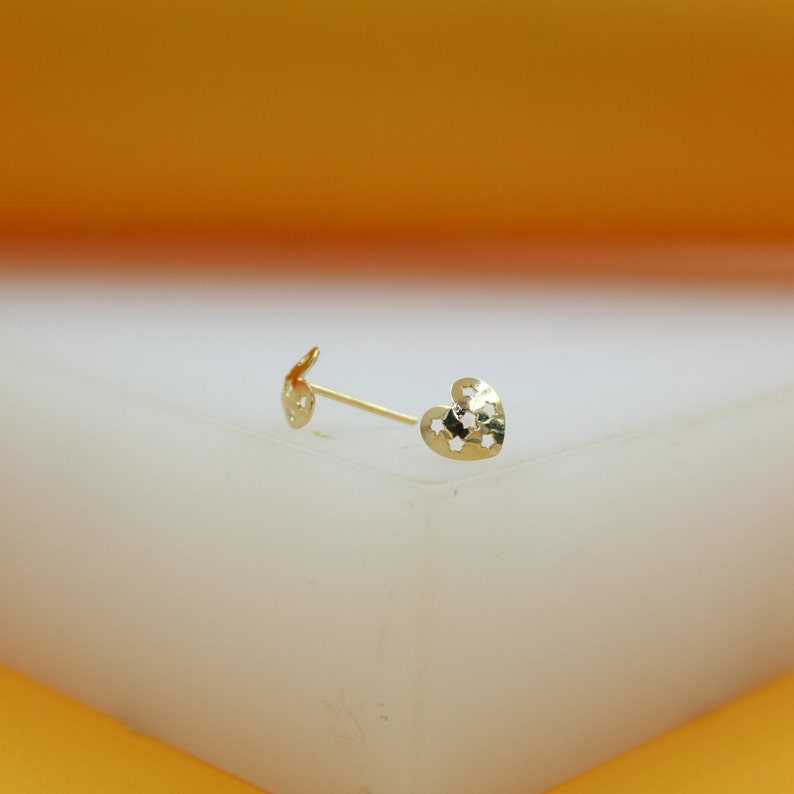 A delicate gold heart stud earring rests on a white display block against an orange backdrop.