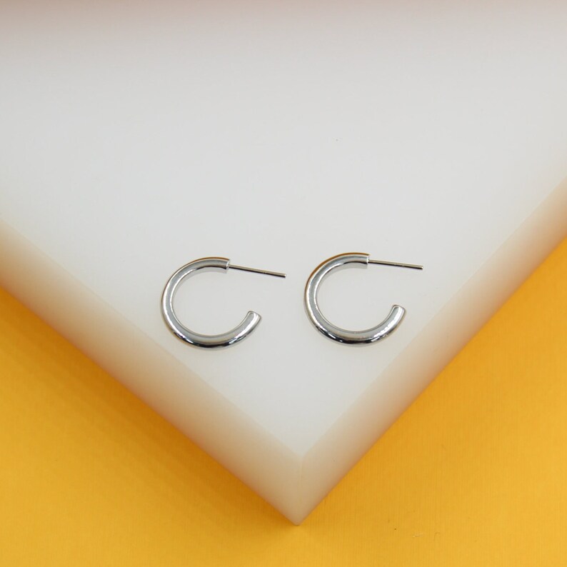 Two polished silver hoop earrings rest on a white display stand against a vibrant yellow background.