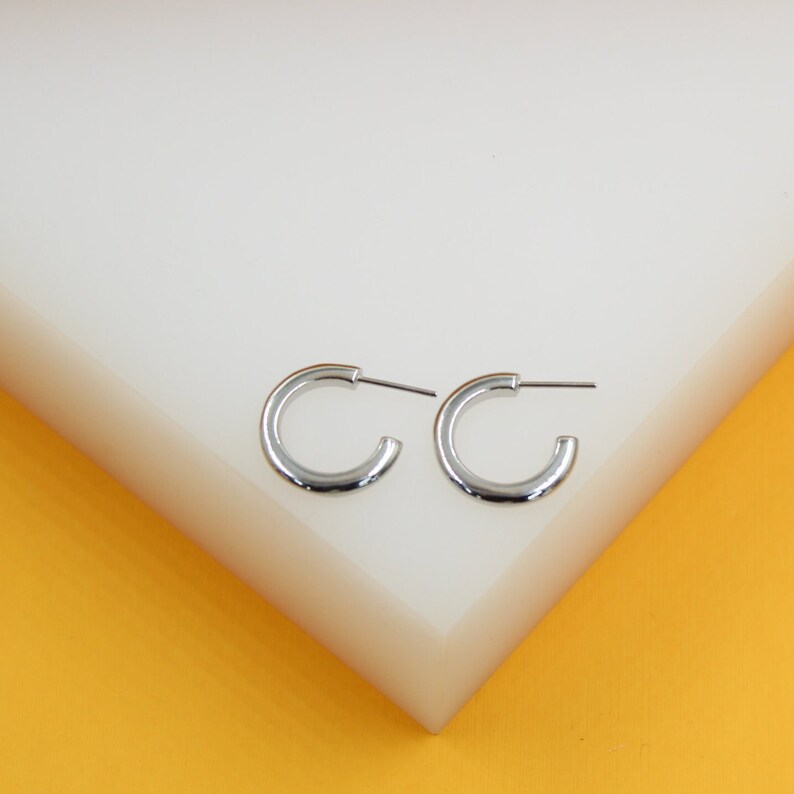 A pair of polished silver hoop earrings rests on a white block against a yellow background.