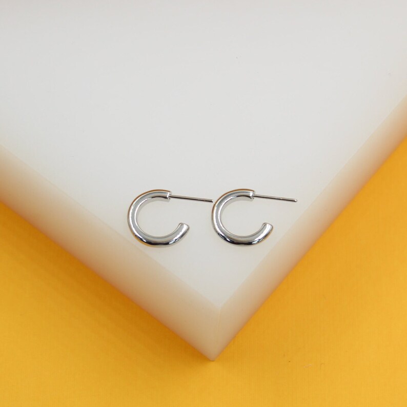 A pair of polished silver half-hoop earrings rests on a white display block against a vibrant yellow background.