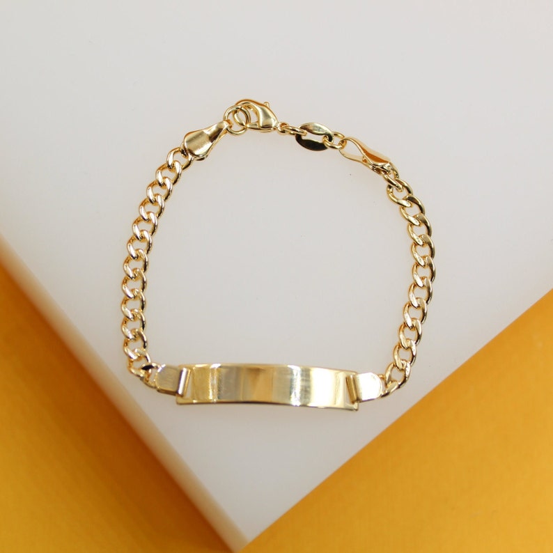 A polished gold ID bracelet with a curb chain rests on a white and yellow surface.