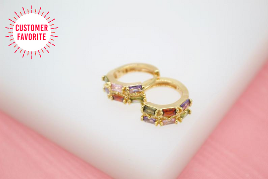 A pair of gold huggie hoop earrings featuring colorful baguette-cut stones rests on a light background next to a "customer favorite" badge.