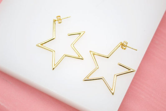 A pair of shiny gold star-shaped hoop earrings rests on a white display surface against a pink background.