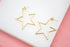 A pair of shiny gold star-shaped hoop earrings rests on a white display surface against a pink background.