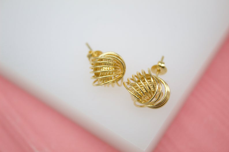 A pair of intricate gold wire ball stud earrings rests on a white and pink display surface.