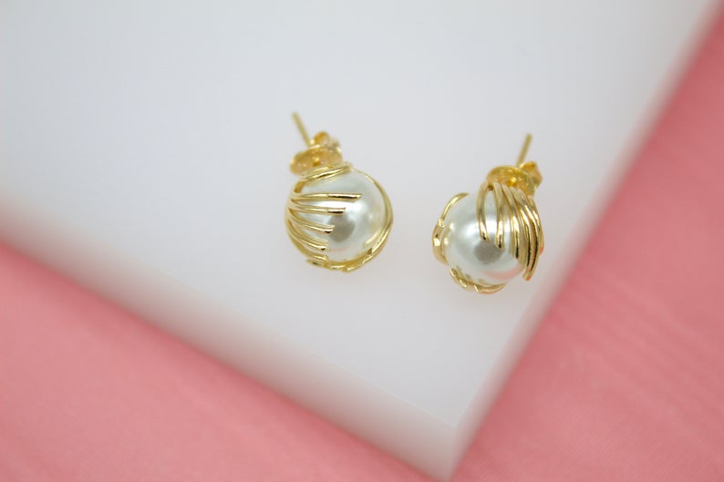 A pair of elegant gold-plated stud earrings featuring white pearls cradled in delicate wirework rest on a white display block.