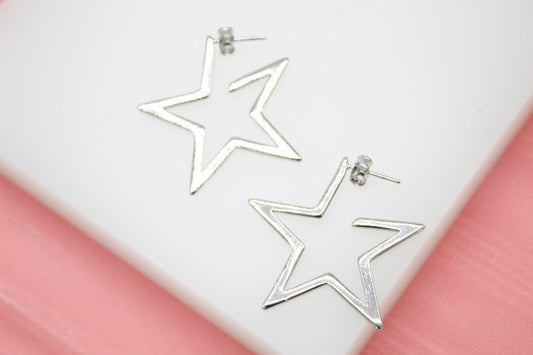 A pair of large silver star-shaped hoop earrings rests on a white display stand against a soft pink background.