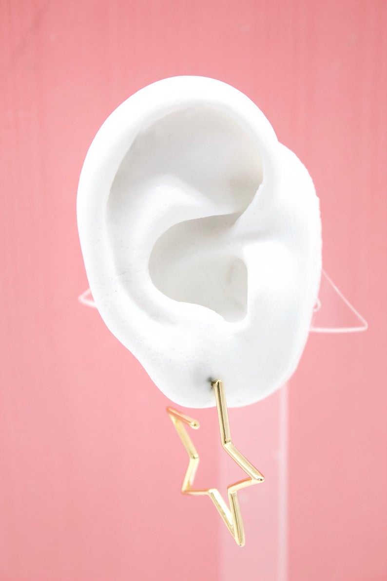 A white mannequin ear displays a shiny gold star-shaped hoop earring against a vibrant pink background.