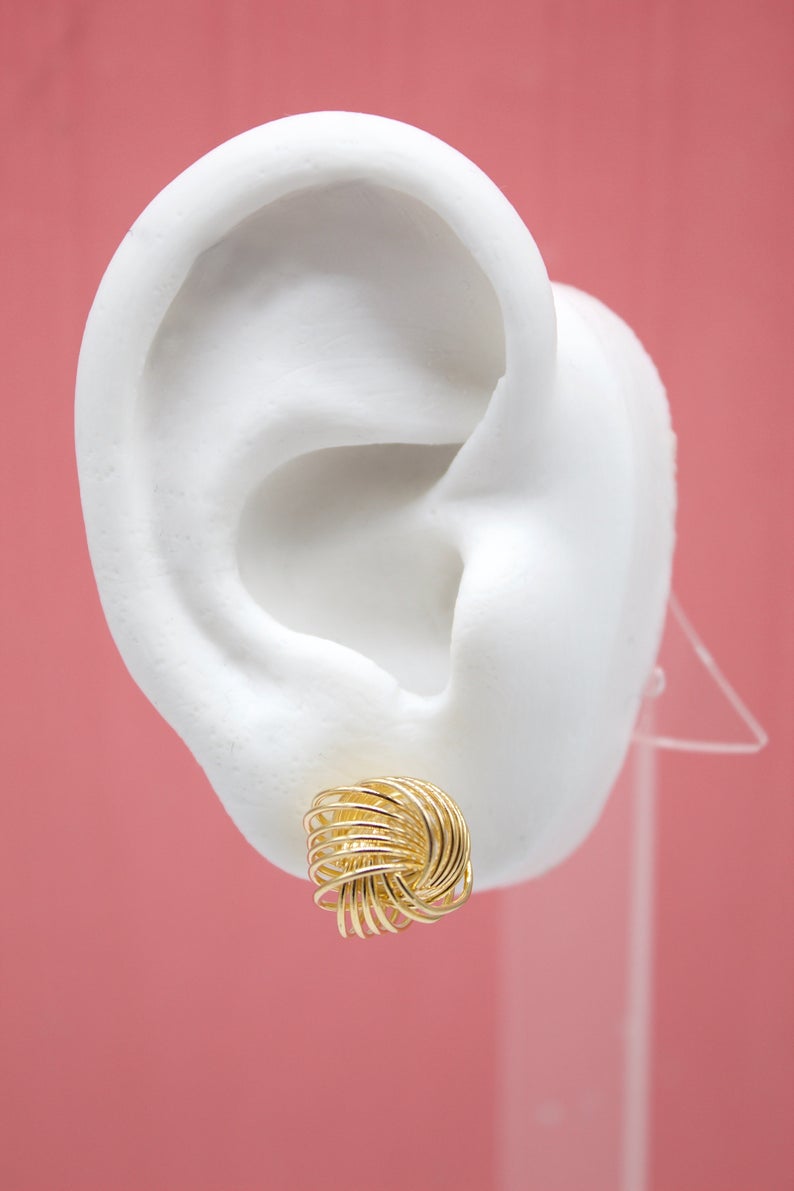 A white display ear model showcases an intricate gold wire knot stud earring against a solid pink background.