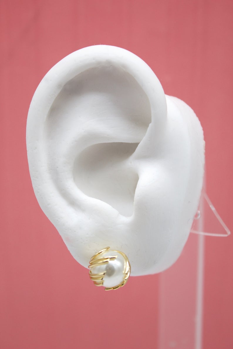 A white mannequin ear displays a gold pearl stud earring against a pink background.