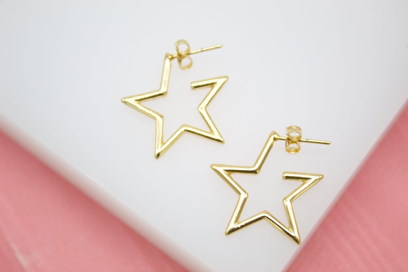 A pair of shiny gold star-shaped hoop earrings rests against a white and pink background.