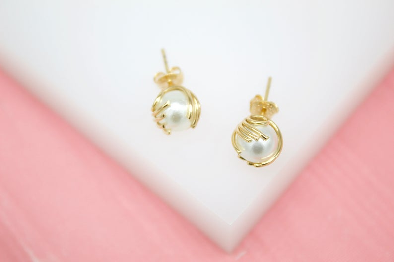 A pair of elegant gold stud earrings features white pearls nestled inside intricate wirework cages resting on a white display surface.