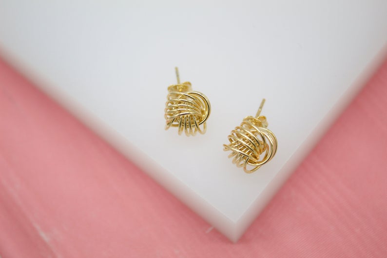 A pair of intricate gold wire-wrapped knot stud earrings rests on a white display against a pink background.