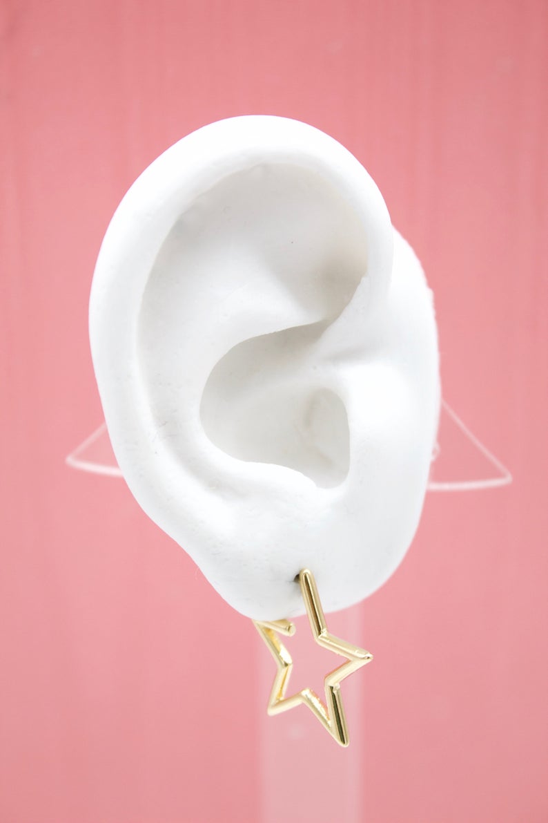 A white mannequin ear displays a gold star-shaped hoop earring against a pink background.