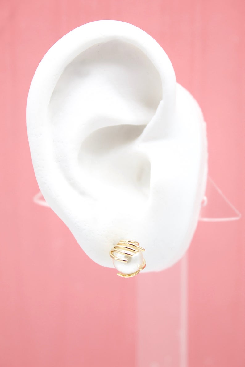 A white mannequin ear displays a gold-wrapped pearl stud earring against a pink background.