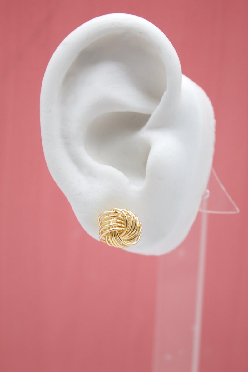 A white mannequin ear displays a textured gold knot stud earring against a pink background.
