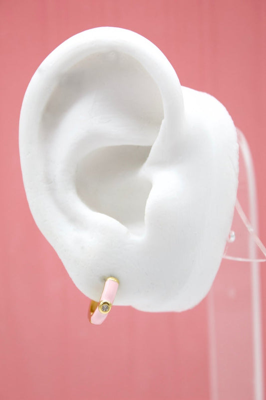 A white mannequin ear displays a pink enamel huggie hoop earring with a small center diamond against a solid pink background.