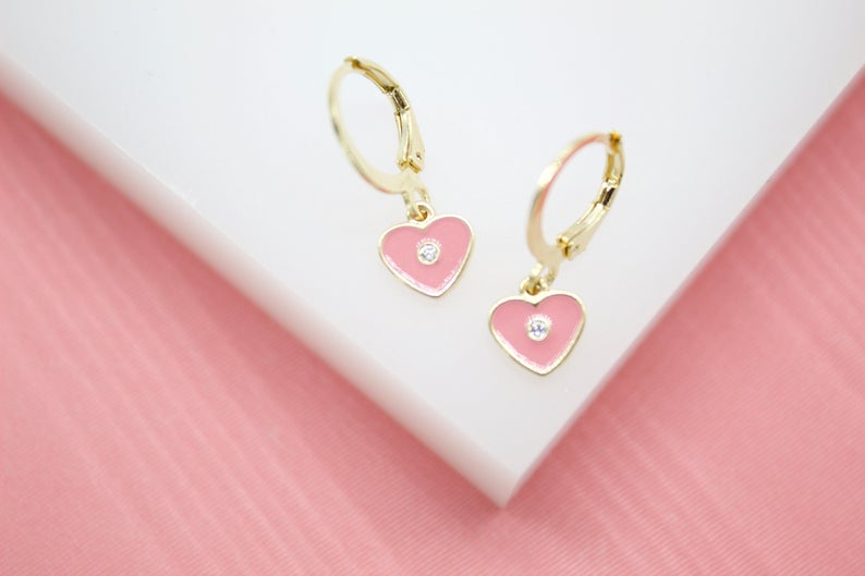 A pair of gold huggie hoop earrings features dangling pink enamel heart charms accented with sparkling CZ stones.