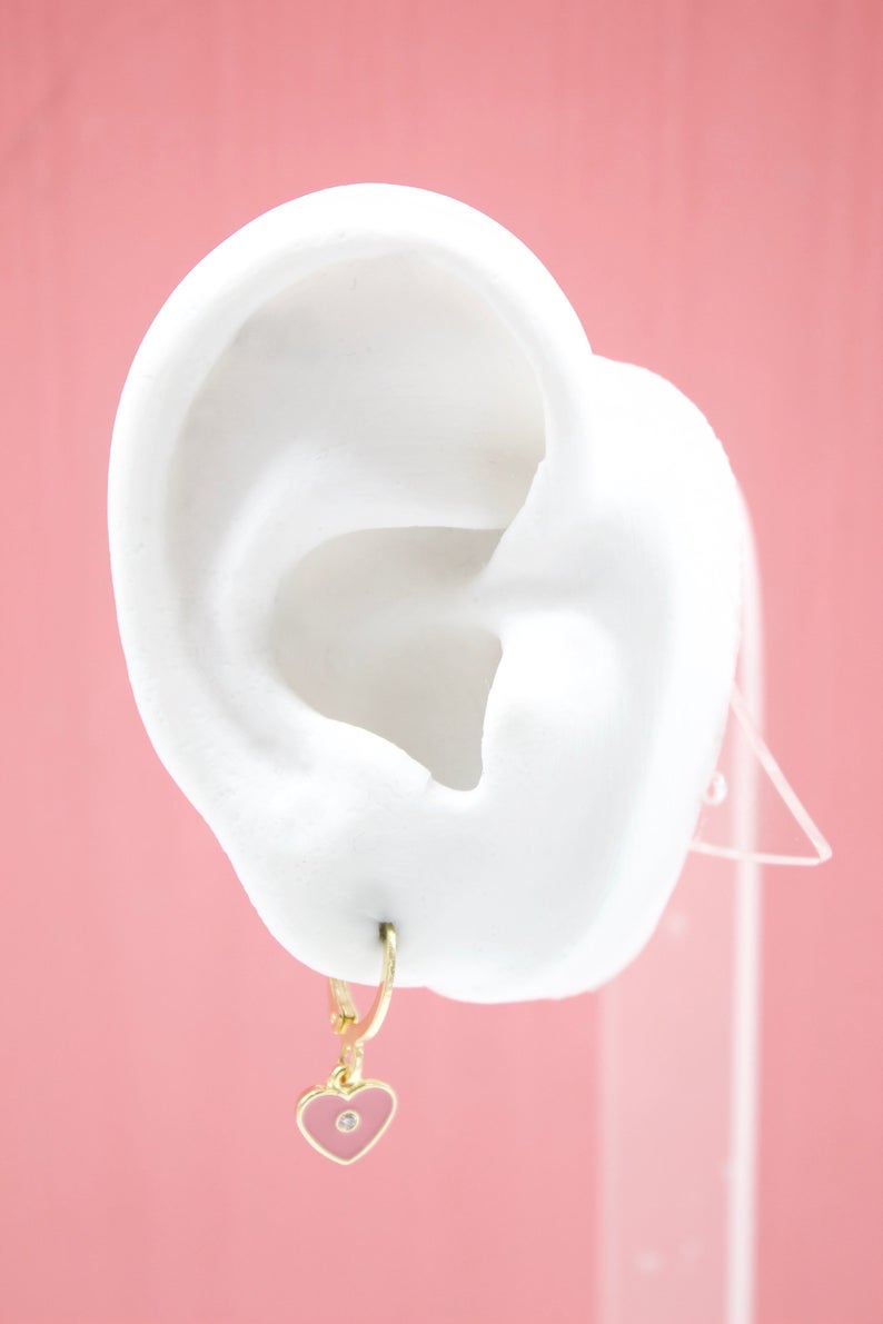 A white mannequin ear displays a gold huggie hoop earring featuring a pink enamel heart charm with a tiny gemstone in the center.