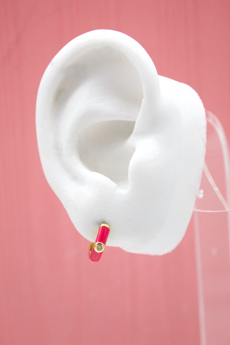 A white mannequin ear displays a vibrant hot pink enamel hoop earring accented with a sparkling diamond.