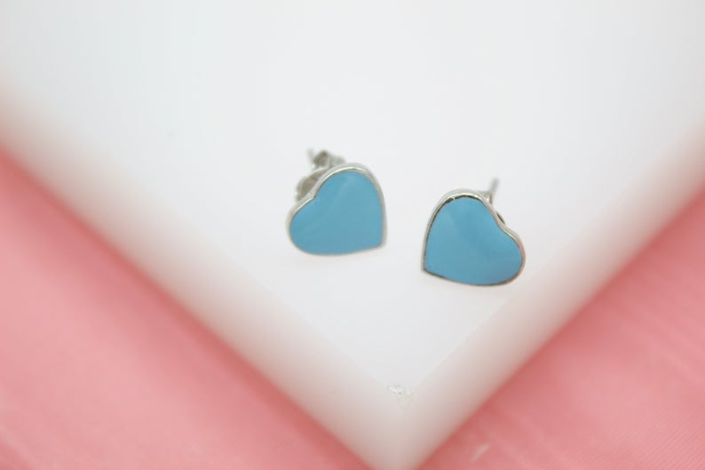 A pair of light blue enamel heart-shaped stud earrings rests on a white display block.