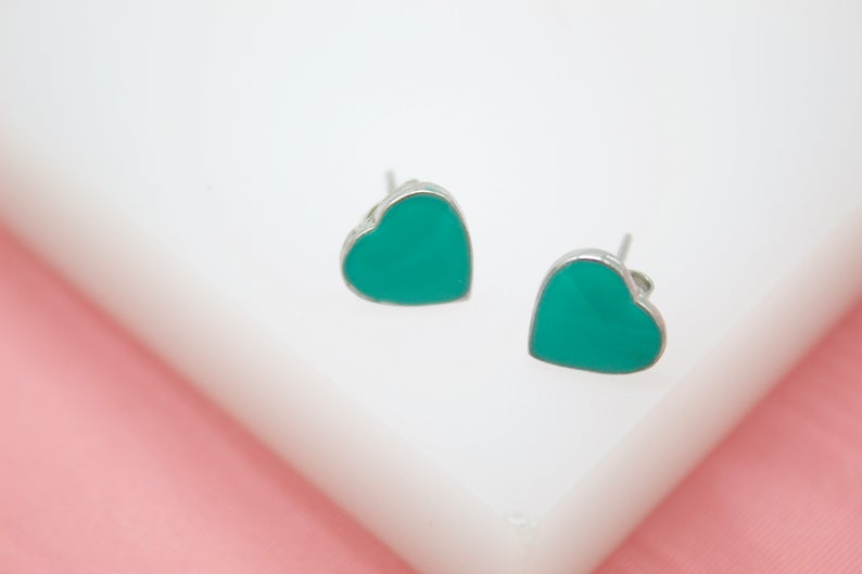 A pair of vibrant green heart-shaped enamel stud earrings rests on a white surface with a pink backdrop.