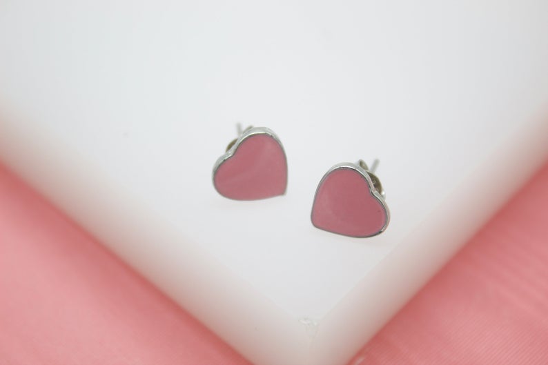 A pair of pink enamel heart stud earrings rests on a white display surface against a pink background.