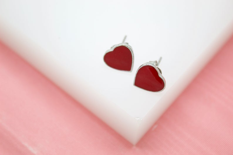 A pair of silver heart-shaped stud earrings with glossy red enamel rests on a white surface.