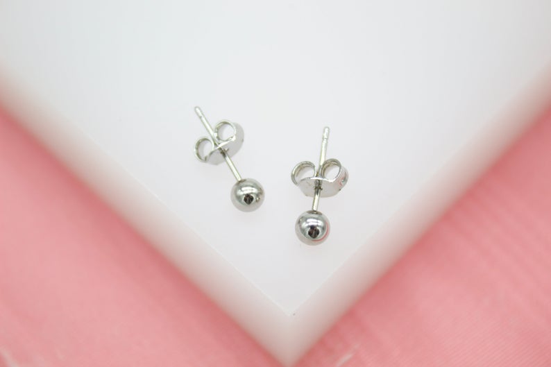 Two simple silver ball stud earrings rest on a white surface against a pink background.