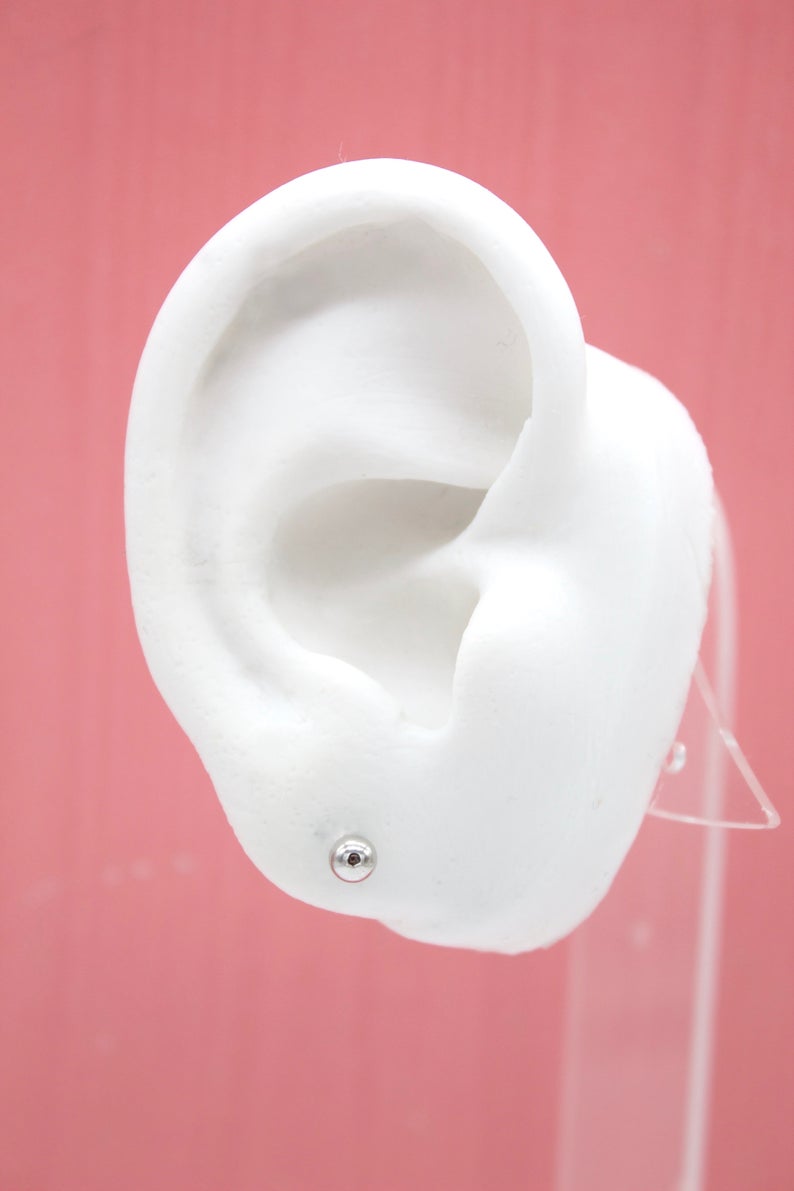 A white mannequin ear displays a small silver ball stud earring against a pink background.