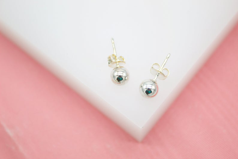 A pair of polished silver ball stud earrings rests on a white display surface against a pink background.