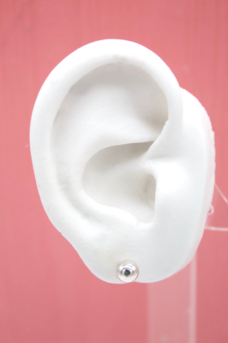 A white anatomical ear model showcases a shiny silver ball stud earring fastened securely on its lobe against a soft pink background.