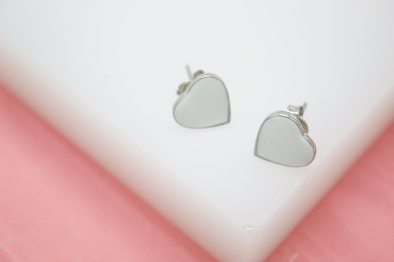 A pair of silver heart-shaped stud earrings rests neatly on a white surface against a soft pink background.