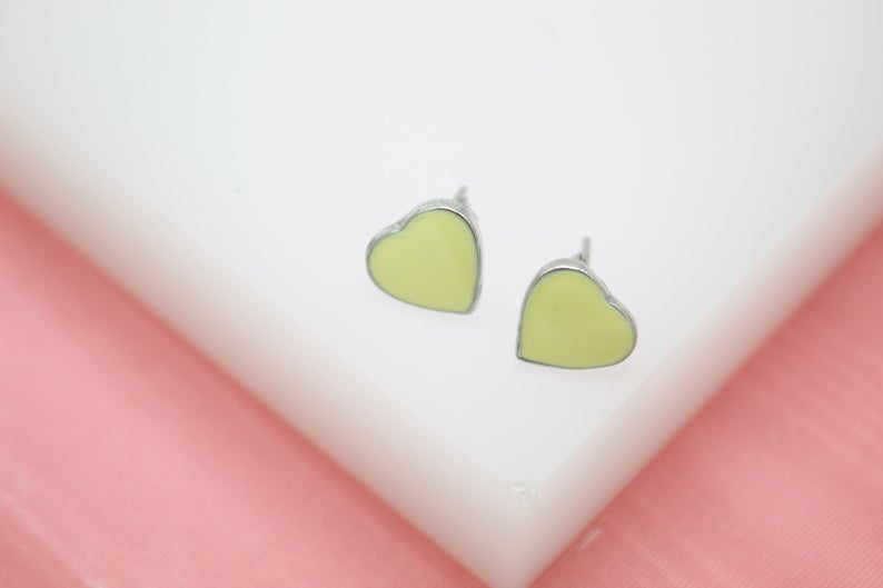 Two light yellow heart-shaped enamel stud earrings rest on a white surface against a pink background.