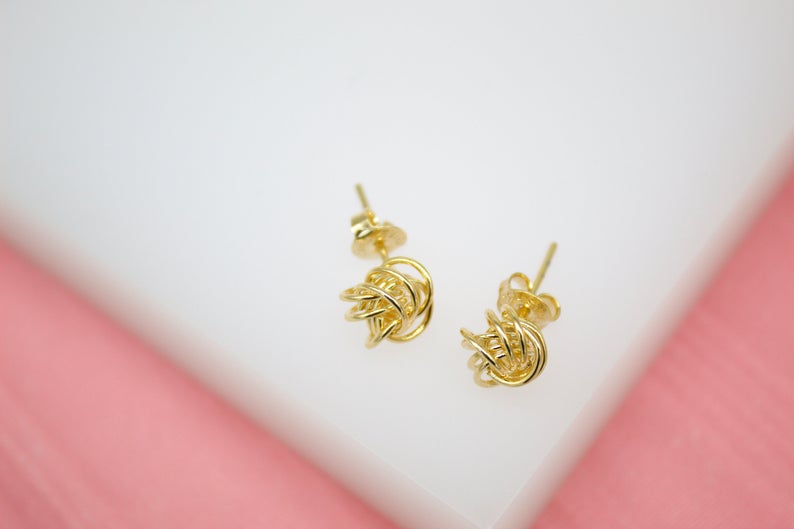 Two intricate gold knot stud earrings rest against a clean white and pink background.