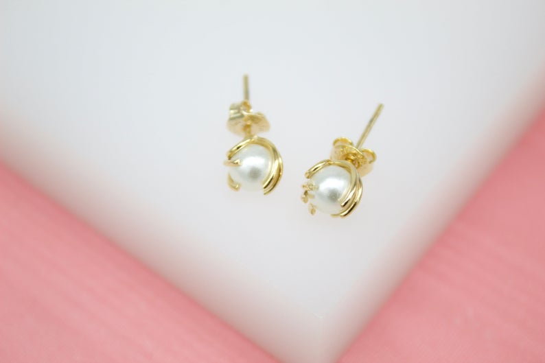 A pair of elegant gold pearl stud earrings rests delicately on a soft white and pink display surface.