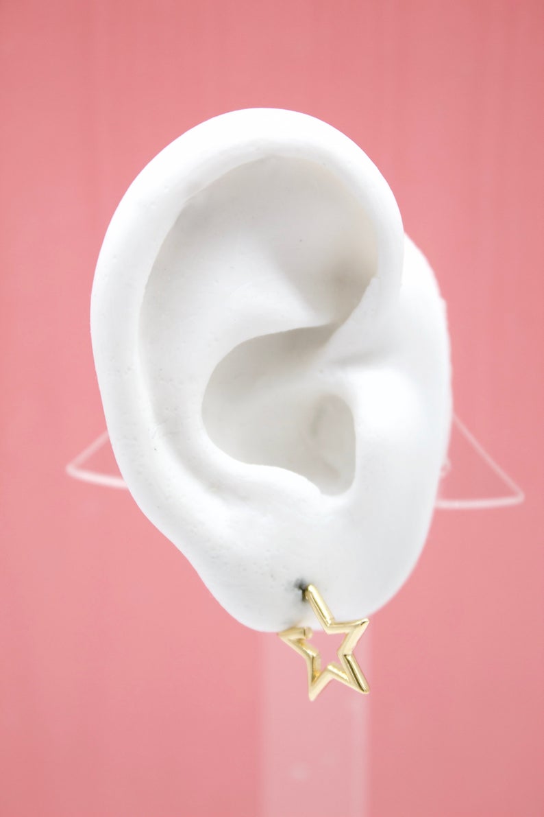 A white model ear displays a shiny gold star-shaped stud earring against a solid pink backdrop.