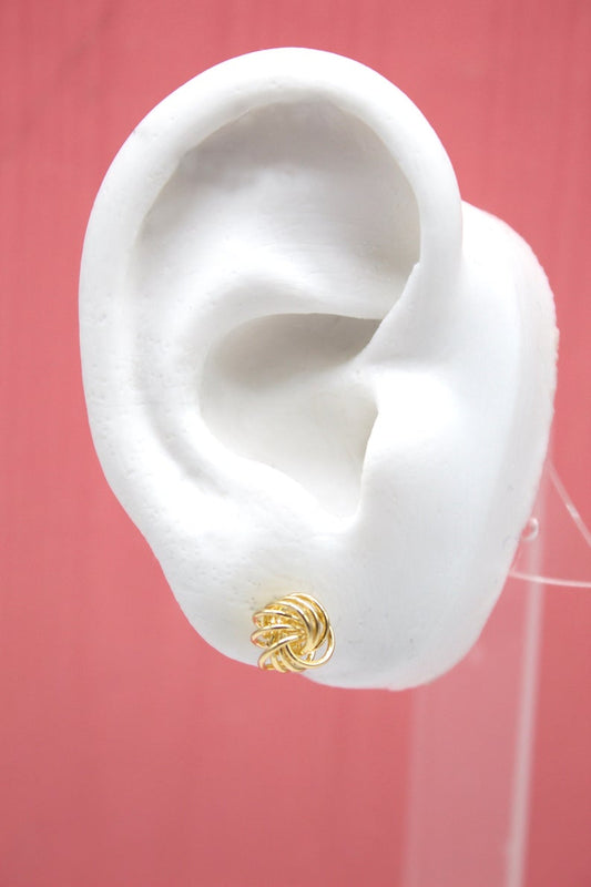 A white model ear showcases a polished gold wire knot stud earring against a solid pink background.