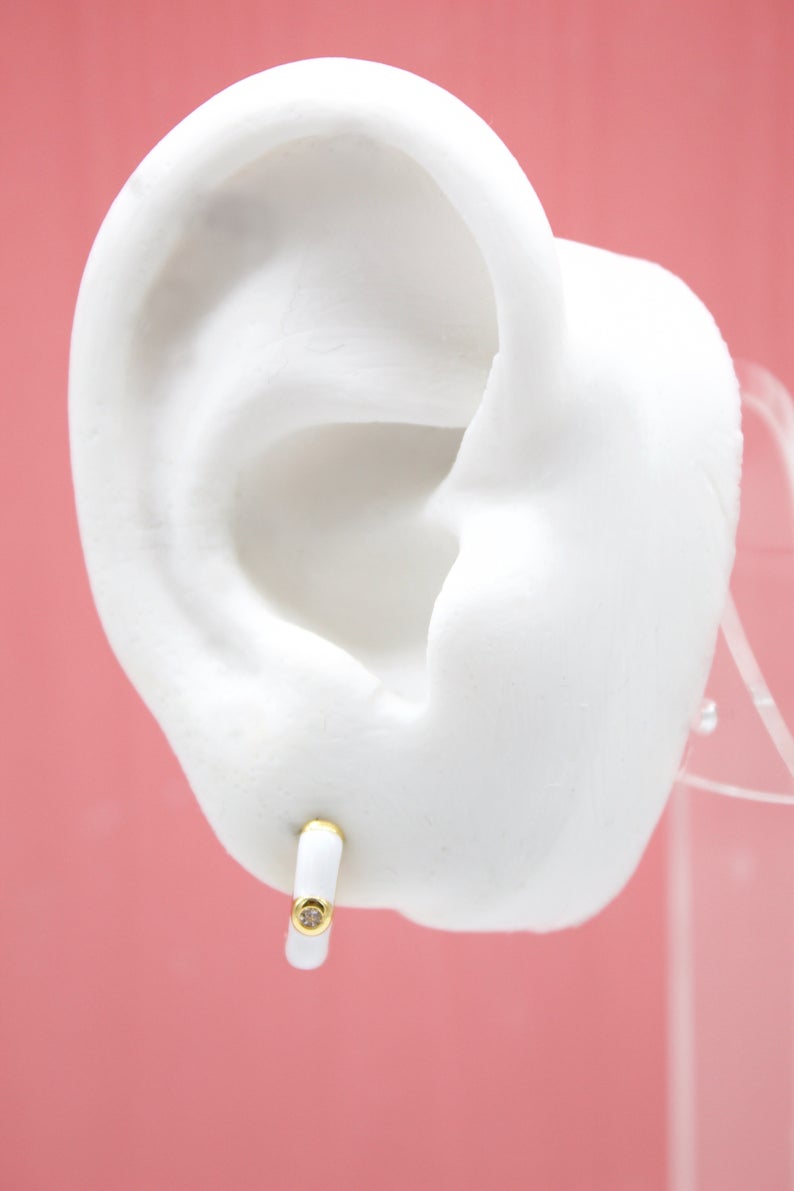 A white mannequin ear displays a gold huggie hoop earring featuring white enamel and a sparkling diamond accent.