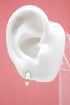 A white mannequin ear displays a gold huggie hoop earring featuring white enamel and a sparkling diamond accent.